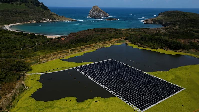 Painel solar flutuante instalado em lago cercado por vegetação verde. Ao fundo, mar azul com ilha rochosa e faixa de areia entre colinas verdes sob céu claro.