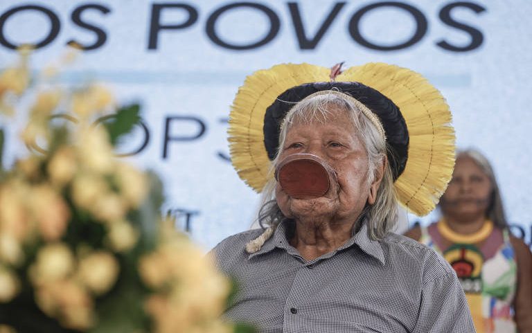 Homem indígena idoso usa cocar amarelo e preto e disco labial grande em evento com fundo de texto parcialmente visível. Outra pessoa indígena aparece desfocada ao fundo.
