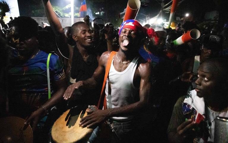 Homens em festa noturna tocam tambores e instrumentos de percussão em ambiente ao ar livre. Um homem no centro usa faixa vermelha na cabeça e camiseta regata branca, expressando energia enquanto toca o tambor.