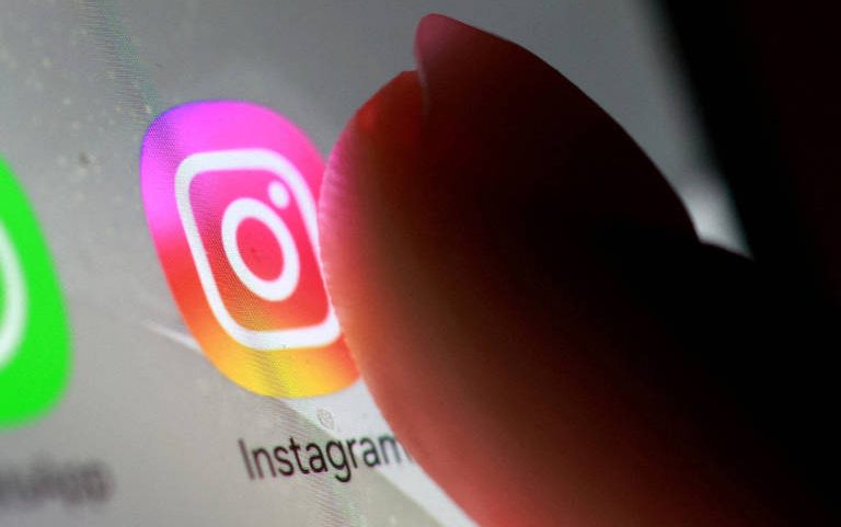 Dedo em foco tocando o ícone colorido do aplicativo Instagram na tela de um smartphone. Ícone apresenta degradê em tons de rosa, roxo, amarelo e laranja com símbolo branco no centro.
