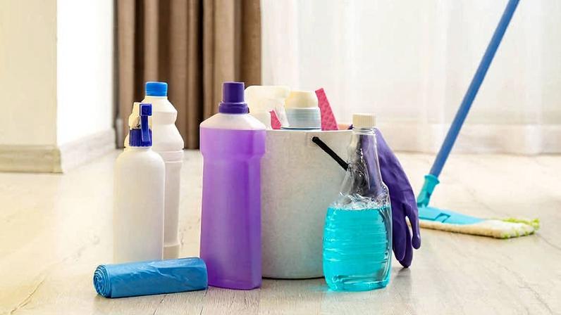 Vários produtos de limpeza, incluindo frascos com líquidos, um rolo de saco plástico azul, luvas roxas e panos, estão organizados próximos a um balde branco. Ao fundo, um mop azul com esfregão verde está apoiado no chão de piso claro, em ambiente interno com cortinas marrons e janela com luz natural.