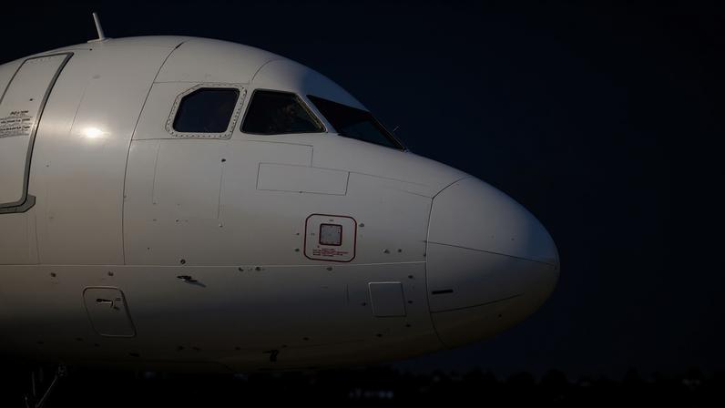 Close do nariz de um avião comercial branco com piloto visível na cabine. Fundo mostra árvores e placas com logotipo da TAM em aeroporto.
