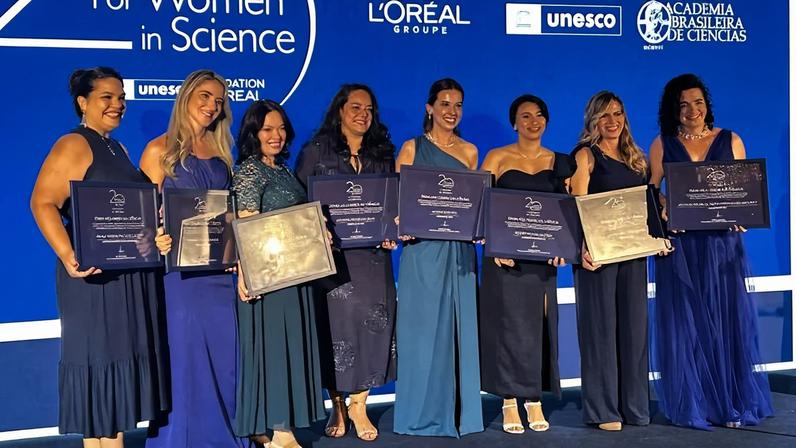 Sete mulheres vestidas com roupas formais em tons de azul posam em palco segurando placas de premiação. Ao fundo, painel azul com logotipos da L'Oréal, Unesco e Academia Brasileira de Ciências, e texto "Para mulheres na ciência em parceria com".