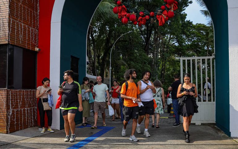Grupo de pessoas caminha sob arco azul e branco decorado com guirlanda vermelha. Ambiente externo com árvores ao fundo e portão branco aberto.