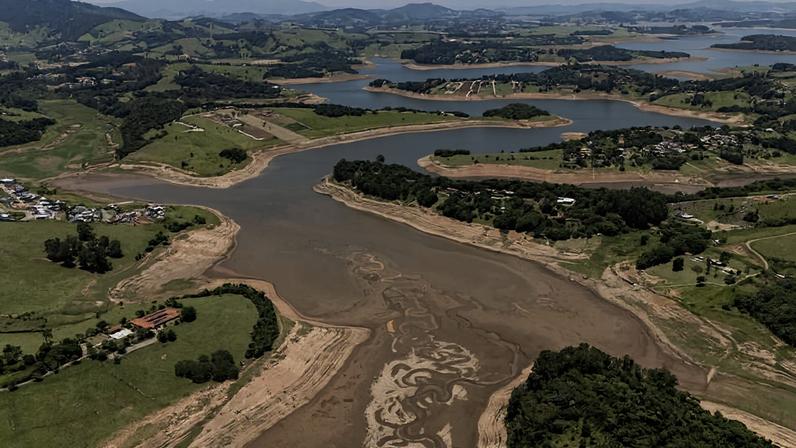 Vista aérea mostra represa com nível de água visivelmente baixo, revelando margens de terra exposta. Áreas verdes e pequenas construções cercam o reservatório, com colinas ao fundo sob céu parcialmente nublado.