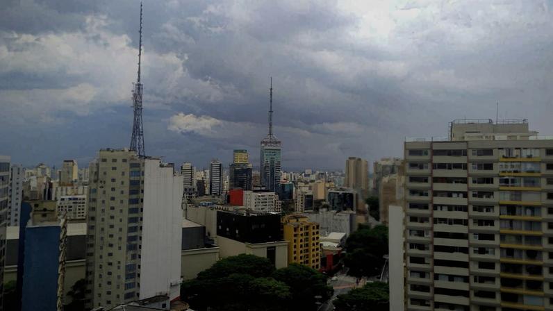 Vista panorâmica da cidade de São Paulo com prédios altos, incluindo a Torre da TV e a Torre do Banespa ao fundo, sob céu nublado com nuvens densas. Áreas verdes e ruas são visíveis entre os edifícios.