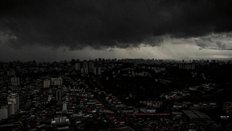 Nuvens escuras e carregadas cobrem uma grande área urbana com prédios residenciais e comerciais. A luz do sol é bloqueada, criando sombras intensas sobre a cidade.