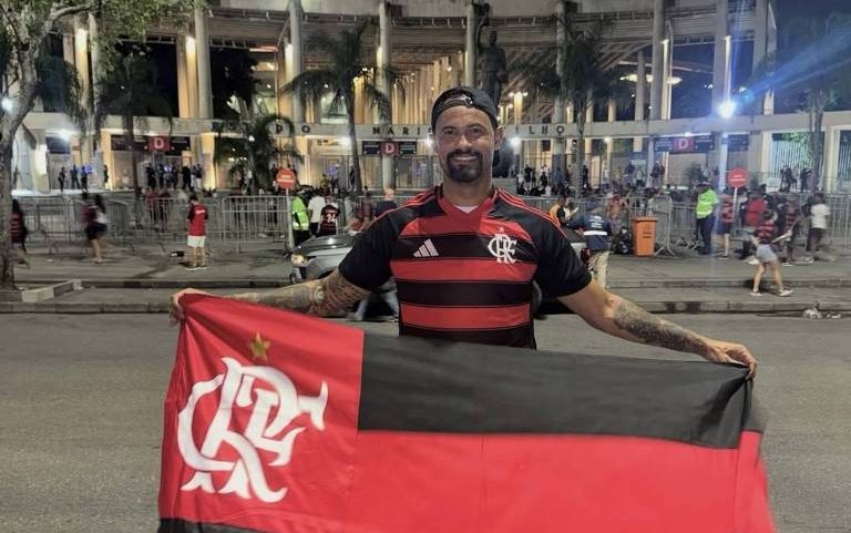 Homem segura bandeira do Flamengo com listras vermelhas e pretas em frente a estádio iluminado à noite. Pessoas e grades estão ao fundo na rua.