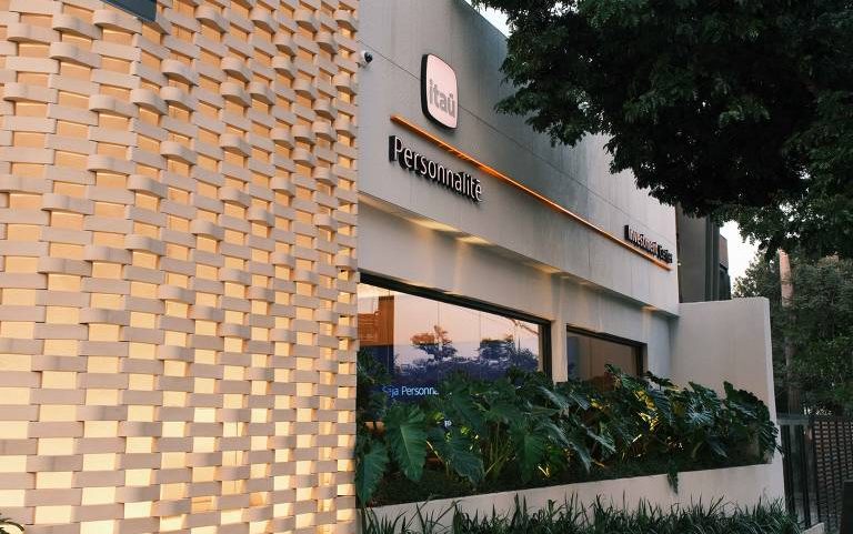 Fachada externa da agência Itaú Personnalité com parede texturizada iluminada à esquerda e vegetação em frente às janelas. Árvore grande à direita e céu ao entardecer refletem na vidraça.