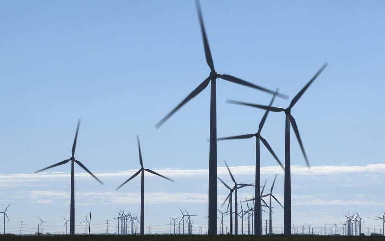 Diversas turbinas eólicas alinhadas em campo aberto sob céu azul claro com poucas nuvens. As pás das turbinas estão em movimento, indicando geração de energia eólica.