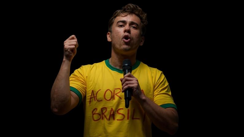 Jovem com camiseta amarela e detalhes verdes segura microfone e gesticula durante discurso em evento ao ar livre. Pessoas e câmeras ao redor capturam o momento.