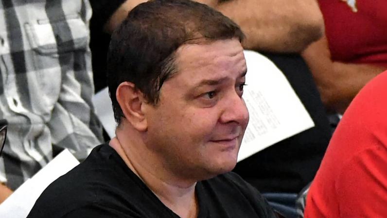 Homem de cabelo curto e escuro veste camiseta preta e está sentado, olhando para a direita. Ao fundo, outras pessoas aparecem parcialmente, uma com camisa xadrez preta e branca e outra com camiseta vermelha.