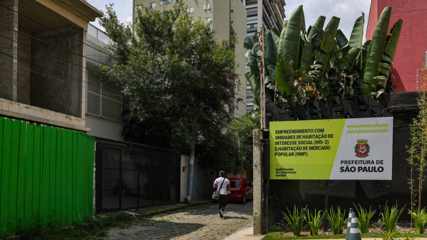 Pessoa caminha por rua de paralelepípedo entre construções urbanas. Placa da Prefeitura de São Paulo anuncia empreendimento com unidades de habitação de interesse social e mercado popular.