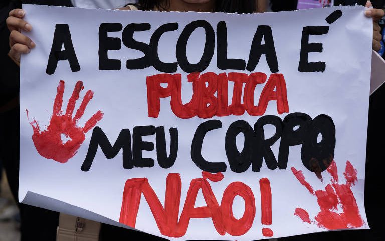 Pessoa segura cartaz com texto "A ESCOLA É PÚBLICA MEU CORPO NÃO!" em letras pretas e vermelhas, com duas marcas de mãos vermelhas ao lado do texto.
