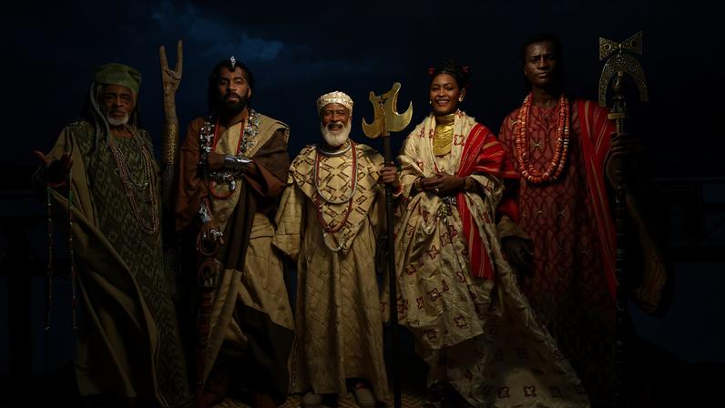 Cinco pessoas vestem roupas tradicionais africanas ornamentadas, com colares grandes e bastões cerimoniais, posando em área externa com céu nublado ao fundo.