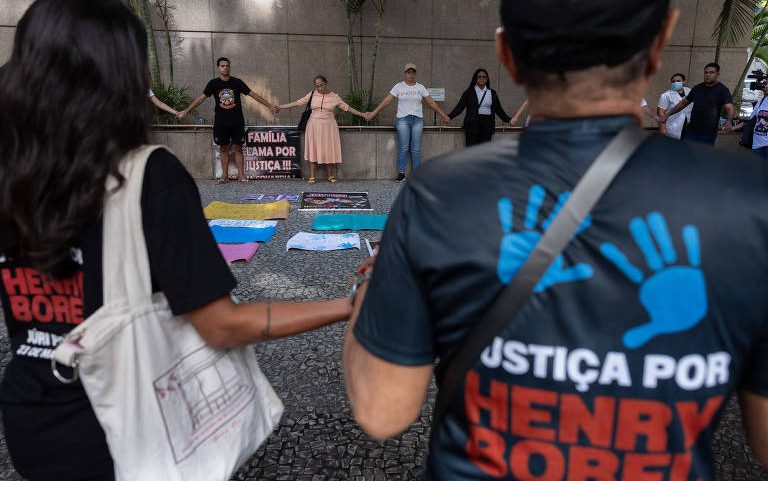 Grupo de pessoas de mãos dadas em protesto contra violência, com cartaz e faixas no chão. Dois manifestantes em primeiro plano usam camisetas com a frase 'Justiça por Henry Borel'.