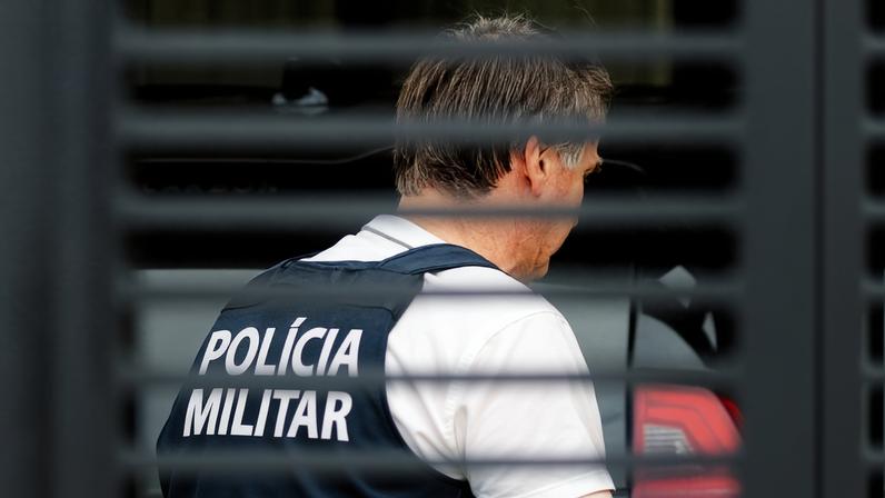 Homem de perfil veste colete preto com a inscrição 'POLÍCIA MILITAR' em branco nas costas. A imagem é capturada através de uma grade ou persiana, desfocando parcialmente a visão. Fundo mostra parte de veículo escuro.