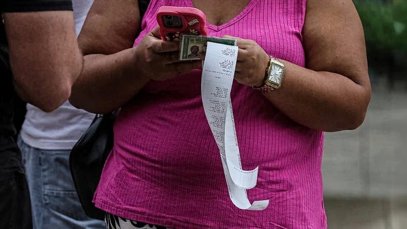 Pessoa com blusa rosa e saia estampada segura celular rosa, dinheiro e um recibo longo pendurado. Outras pessoas aparecem parcialmente ao fundo.