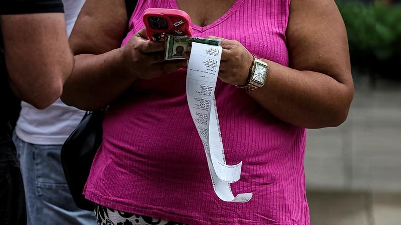 Pessoa com blusa rosa e saia estampada segura celular rosa, dinheiro e um recibo longo pendurado. Outras pessoas aparecem parcialmente ao fundo.