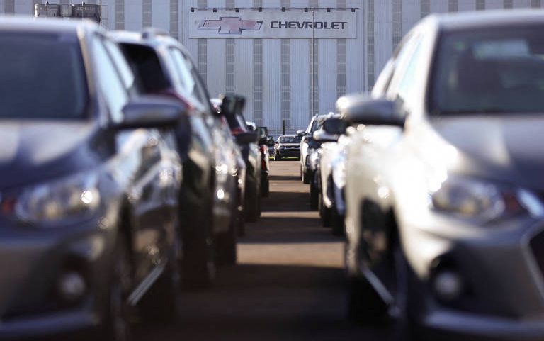 Várias fileiras de carros estacionados lado a lado em área externa, com prédio industrial ao fundo exibindo logotipo e nome Chevrolet.