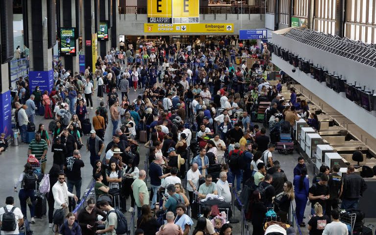 Multidão de passageiros aguarda em filas para check-in em terminal de aeroporto. Painéis indicam setor E e área de embarque, com balcões de atendimento à direita e pessoas com bagagens diversas.