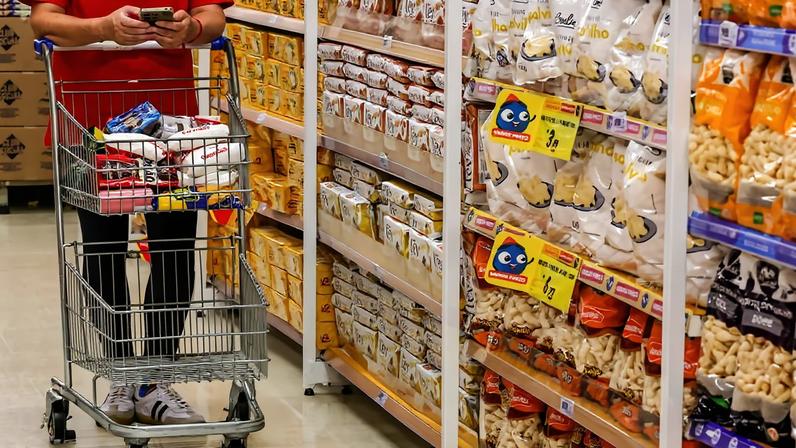 Pessoa vestindo camiseta vermelha e calça preta empurra carrinho de compras em corredor de supermercado. Prateleiras exibem diversos pacotes de biscoitos e salgadinhos.