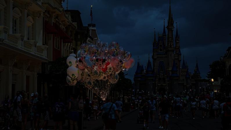 Pessoas caminham e se reúnem na Main Street dos parques Disney, com vendedor carregando balões coloridos de personagens. Ao fundo, o castelo azul e rosa sob céu parcialmente nublado.