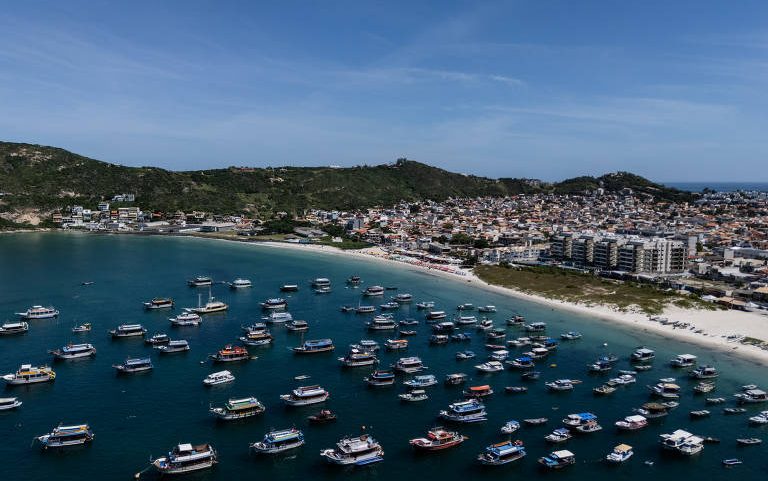 Vista aérea mostra dezenas de barcos ancorados em águas calmas próximas a uma praia extensa. Ao fundo, há uma cidade com prédios residenciais e colinas verdes sob céu azul claro.