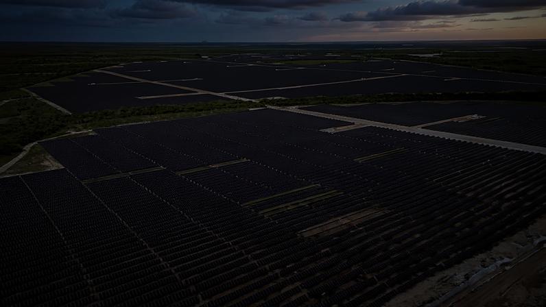 Vista aérea mostra grande usina solar com milhares de painéis fotovoltaicos organizados em fileiras retas sobre terreno plano, cercada por vegetação e céu parcialmente nublado ao fundo.