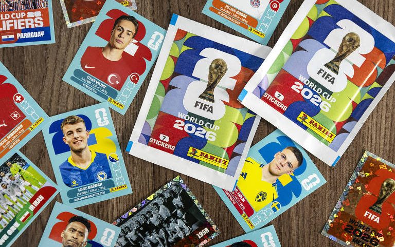 Pacotes e figurinhas soltas da coleção oficial da Copa do Mundo FIFA 2026 estão distribuídos sobre uma superfície de madeira. As figurinhas mostram jogadores de diferentes seleções, incluindo Turquia, Suécia e Suíça, com seus uniformes e bandeiras nacionais. Dois pacotes fechados da coleção oficial aparecem ao centro, com o logo da FIFA e o troféu da Copa do Mundo.
