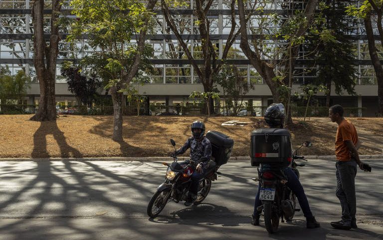 Dois motociclistas parados em rua sombreada por árvores, um deles conversa com um pedestre à direita. Ao fundo, prédio moderno com fachada de vidro e árvores na calçada.