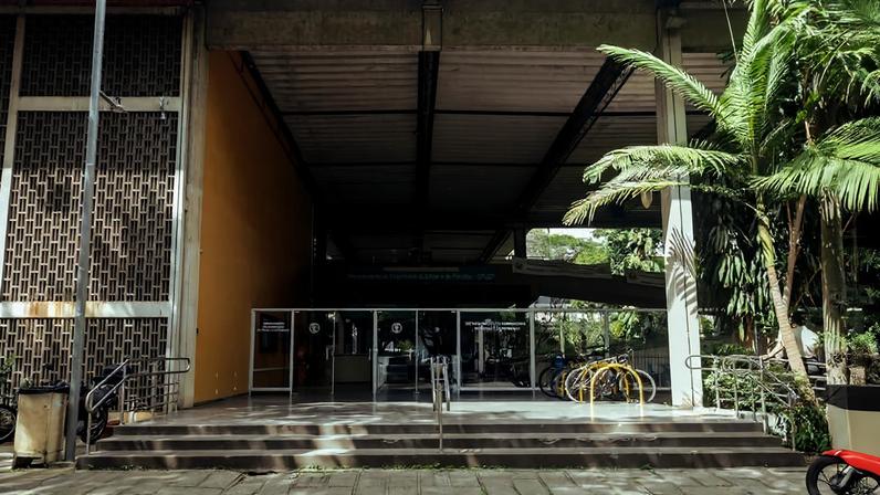 Entrada principal de prédio com escadas e rampa de acesso, portas de vidro automáticas, bicicletário amarelo à direita e vegetação ao redor.