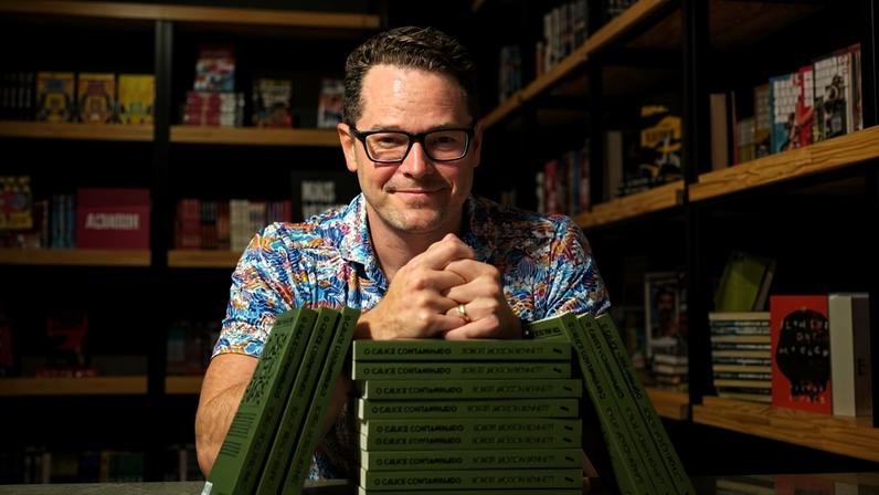 Homem de óculos e camisa estampada está atrás de mesa com pilha organizada de livros verdes intitulados 'O Caixe Contaminado'. Estantes com mais livros ao fundo.