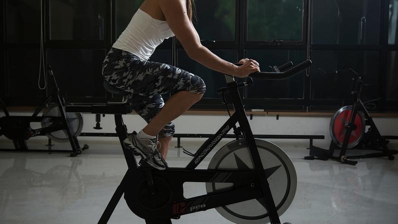 Mulher vestindo regata branca e calça legging camuflada pedala em bicicleta ergométrica preta dentro de academia com piso claro e janelas grandes ao fundo.