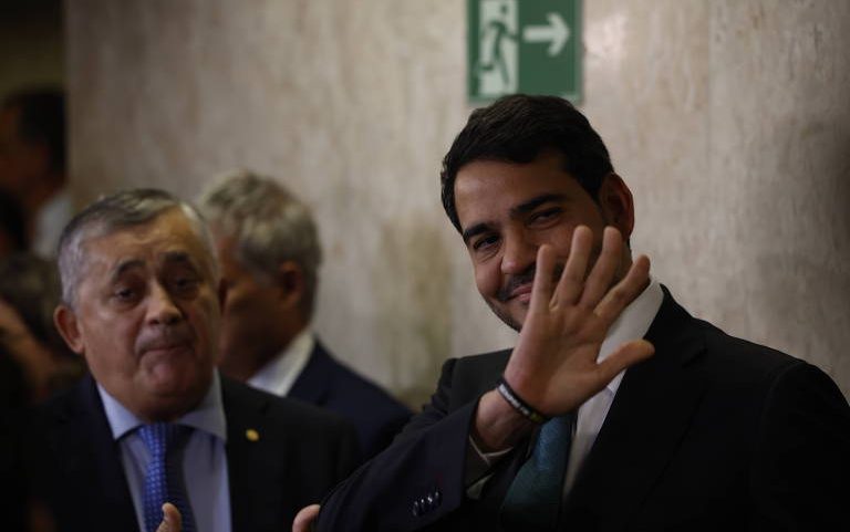 Homem de terno escuro sorri e acena com a mão direita em primeiro plano, com dois homens mais velhos ao fundo, todos em ambiente interno com parede clara e placa de saída de emergência.