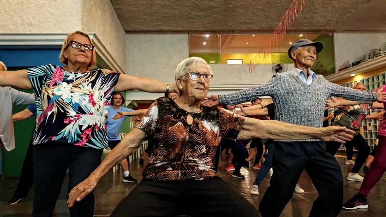 Idosos em sala ampla realizam alongamento com braços abertos e pernas flexionadas. Ambiente tem paredes claras e iluminação artificial.