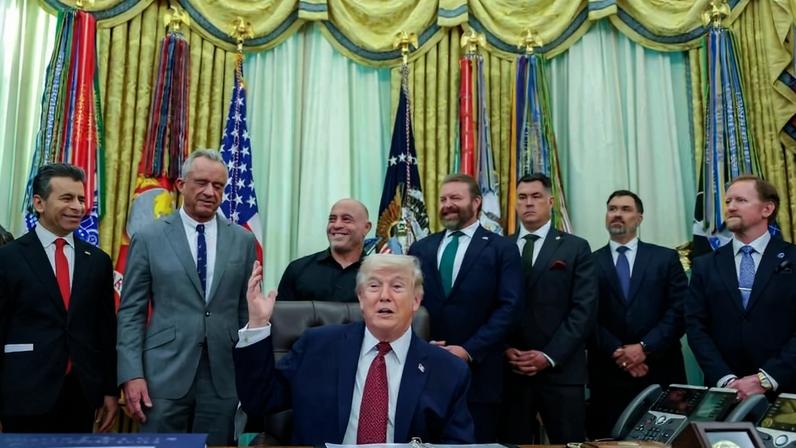 Ex-presidente dos EUA sentado à mesa do Salão Oval, gesticulando com a mão direita levantada. Sete homens em pé atrás dele, todos vestidos formalmente, alguns sorrindo. Bandeiras americanas e decorativas ao fundo, cortinas douradas e iluminação natural pela janela.
