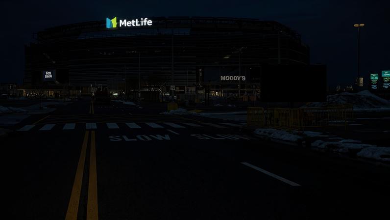Vista frontal do MetLife Stadium com céu nublado. Estacionamento vazio com faixas de pedestres e sinalização 'SLOW' no asfalto. Neve acumulada nas laterais e barreiras amarelas próximas à calçada.