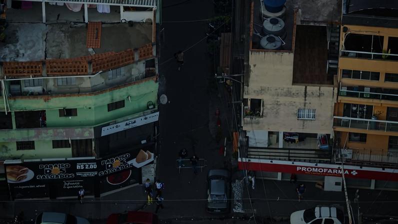 Vista aérea mostra rua entre prédios comerciais com fachadas coloridas. Três carros circulam na via principal, e pedestres caminham nas calçadas. Fachada vermelha de farmácia destaca-se à direita.