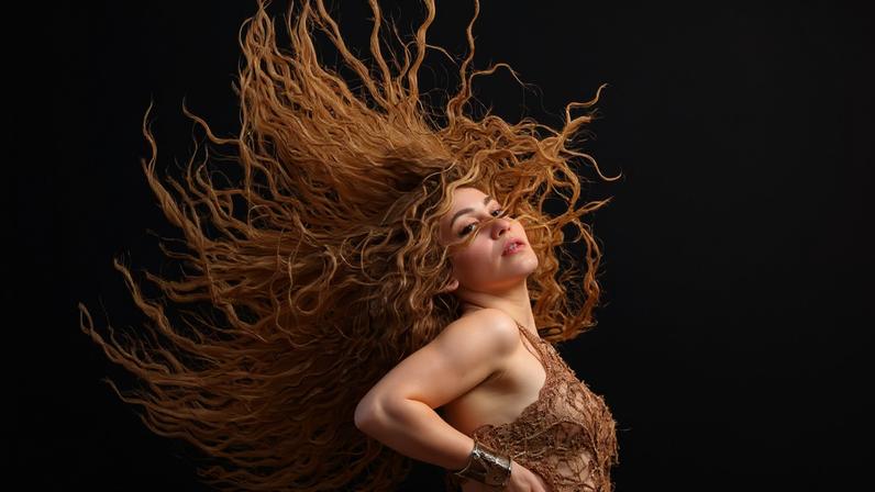 Mulher com cabelo ruivo cacheado esvoaçando em movimento amplo contra fundo preto. Ela veste roupa dourada e está com uma mão na cintura, olhando para frente.