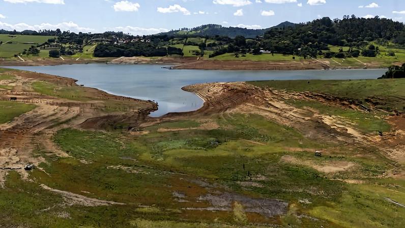 Represa com pouca água visível, cercada por terra seca e áreas de vegetação esparsa. Colinas verdes e árvores ao fundo sob céu azul com poucas nuvens.