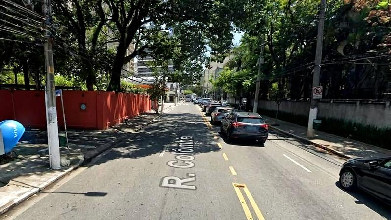 Rua com faixa dupla amarela no centro e carros estacionados no lado direito. Árvores grandes fazem sombra sobre a via. À esquerda, muro vermelho e calçada com poste e orelhão azul. Ambiente urbano durante o dia.