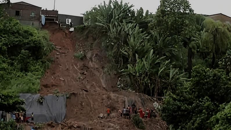 Deslizamento de terra atingiu encosta próxima a casas. Equipes e moradores estão na base da área afetada, cercada por vegetação densa e árvores.