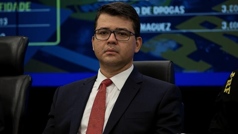 Homem jovem com cabelo curto e óculos, vestido com terno azul escuro, camisa branca e gravata vermelha, sentado em cadeira preta em ambiente institucional com tela azul ao fundo.