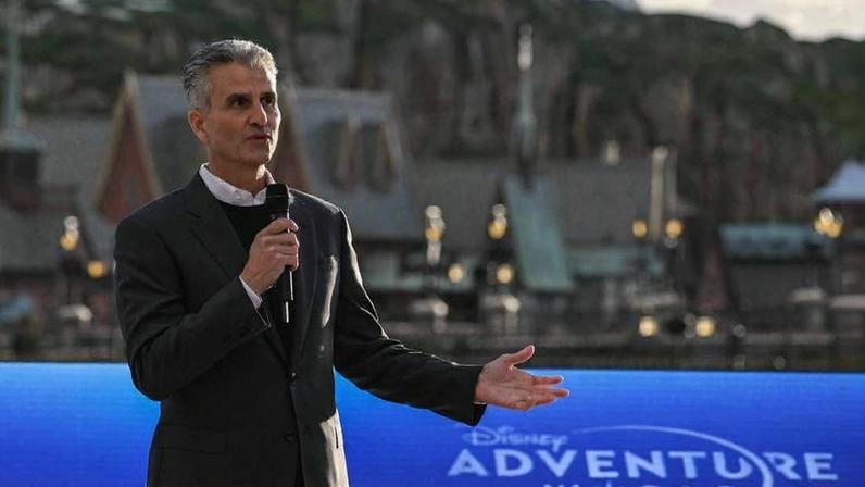 Homem de terno preto segura microfone e gesticula com a mão direita em frente a tela azul com logo da Disney Adventure World. Fundo mostra estruturas e montanhas artificiais do parque.