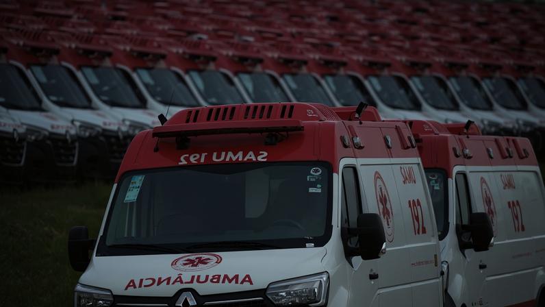 Diversas ambulâncias brancas com teto vermelho estão alinhadas em fileiras em um campo aberto. A ambulância em primeiro plano exibe o texto 'SAMU 192' e o símbolo da cruz vermelha na lateral e na frente.
