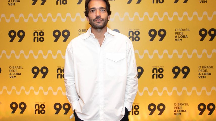 Homem de cabelos escuros e barba curta veste camisa branca e calça preta, posando com as mãos nos bolsos diante de fundo amarelo com logos '99', 'norio' e texto 'O Brasil pede, a Loba vem'. Humberto Carrão chega para assistir ao show de Shakira em Copacabana