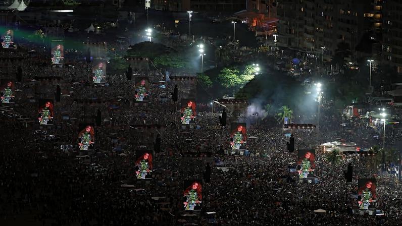 Milhares de pessoas ocupam a praia de Copacabana à noite, diante de vários telões iluminados com imagens coloridas. Luzes e estruturas de som estão distribuídas ao longo da multidão, com prédios ao fundo.