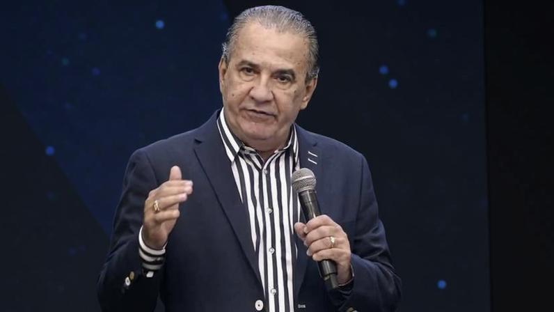 Homem de meia-idade com cabelo grisalho preso para trás, veste paletó azul escuro e camisa listrada preta e branca, fala segurando microfone em púlpito transparente, fundo azul com padrão pixelado.