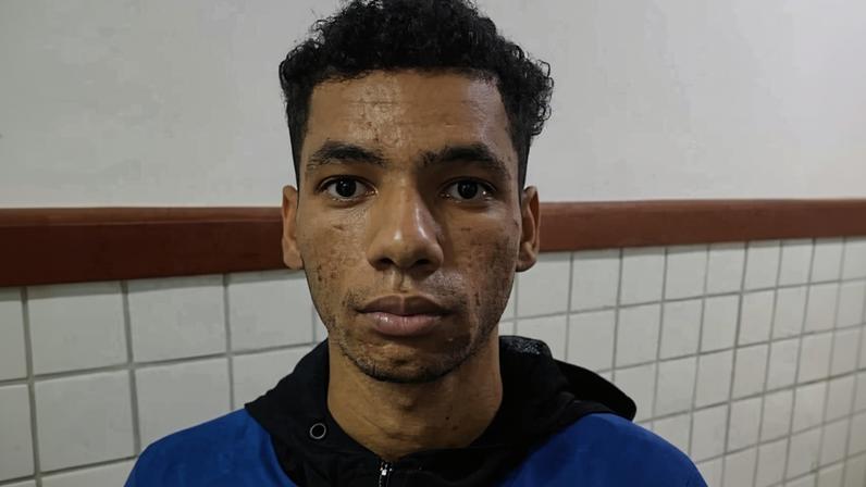 Jovem de pele morena clara, cabelo curto e encaracolado, veste jaqueta azul e preta com logo da Nike. Fundo com parede branca e azulejos brancos com faixa marrom.